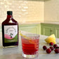 Bottle of Urban Pharm shrub with a glass of red drink and fruit on a kitchen counter.
