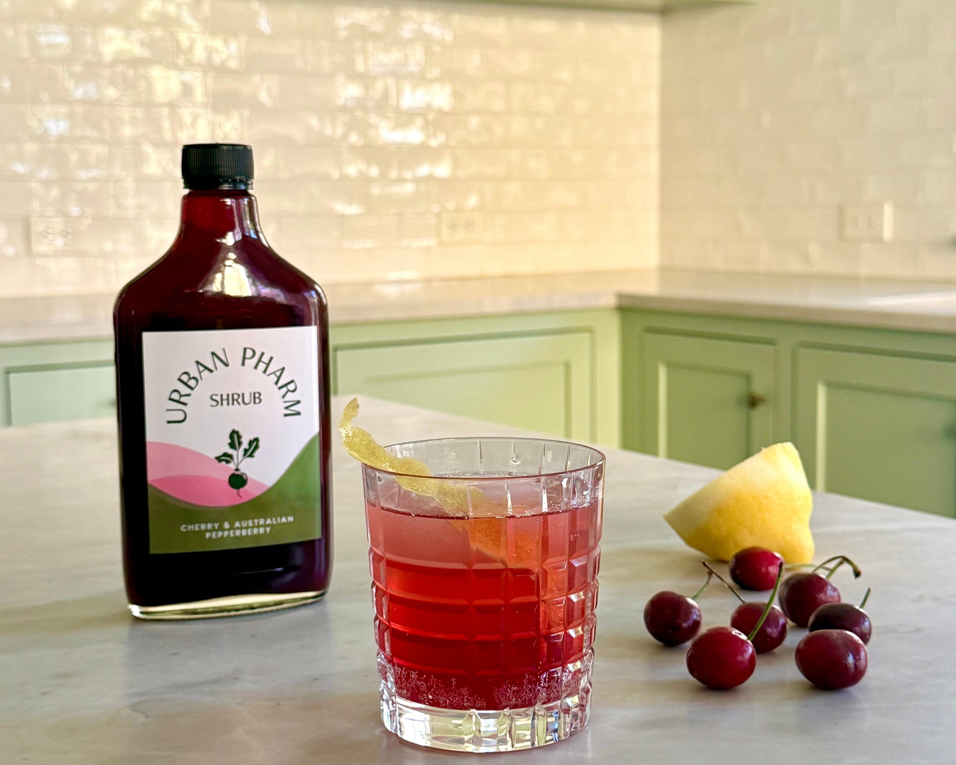 Bottle of Urban Pharm shrub with a glass of red drink and fruit on a kitchen counter.